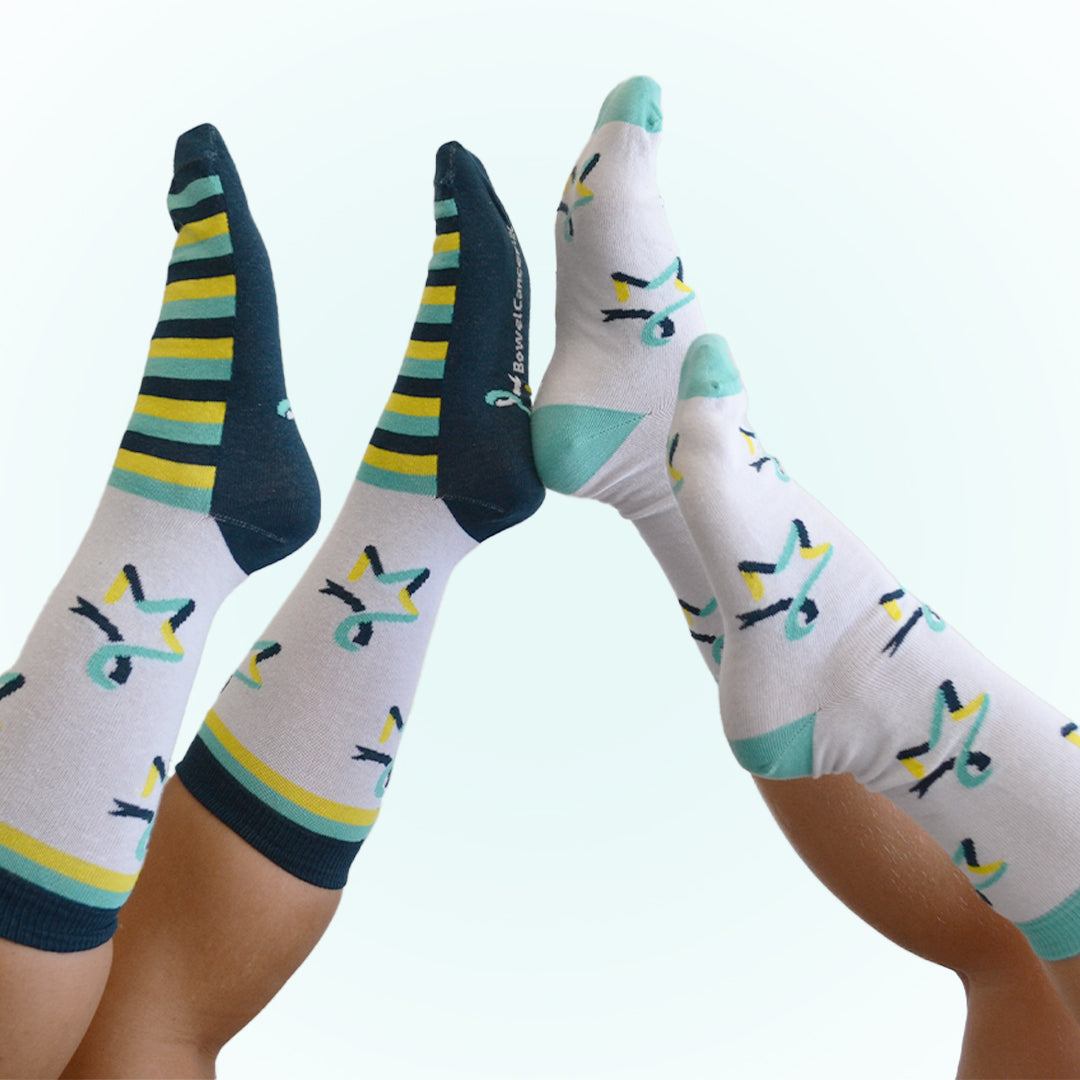 Two pairs of legs are shown from the knees down, angled toward each other with feet pointing upward, both wearing our Star of Hope socks. The pair on the left is wearing white socks with the Star of Hope logo and dark‑teal, yellow, and light‑teal stripes around the ankle cuffs and feet. The pair on the right is wearing white socks with the Star of Hope logo running vertically down the sides, with light‑teal toes and ankle cuffs.