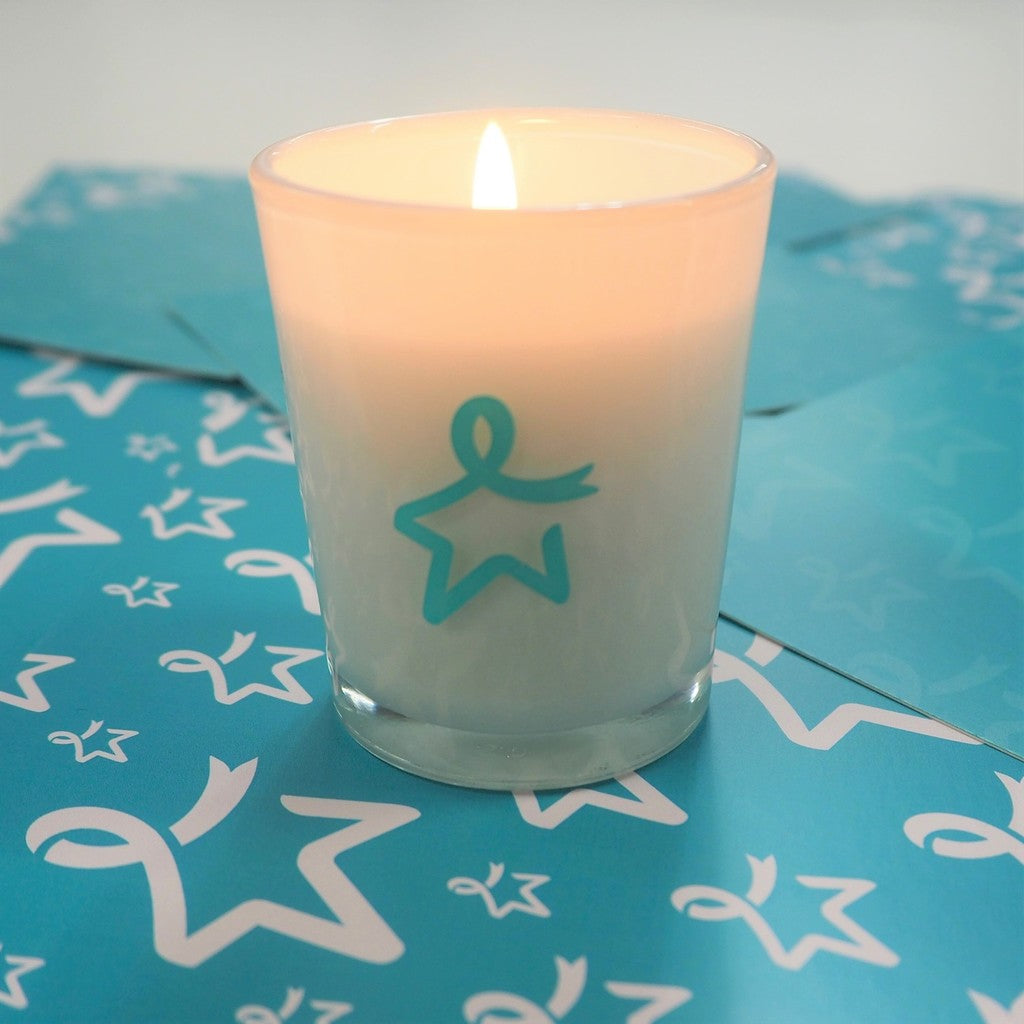 Lit white glass candle with a teal Star of Hope design, displayed on a light blue surface patterned with a white Star of Hope motif.