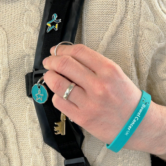 A close-up of a model’s left hand holding a set of keys with a teal trolley coin engraved with the Star of Hope. The model wears a teal wristband on their wrist featuring a multicoloured Star of Hope and the words “Bowel Cancer UK” in white. A black bag strap is also visible, with a pin badge displaying the multicoloured Star of Hope attached to it.