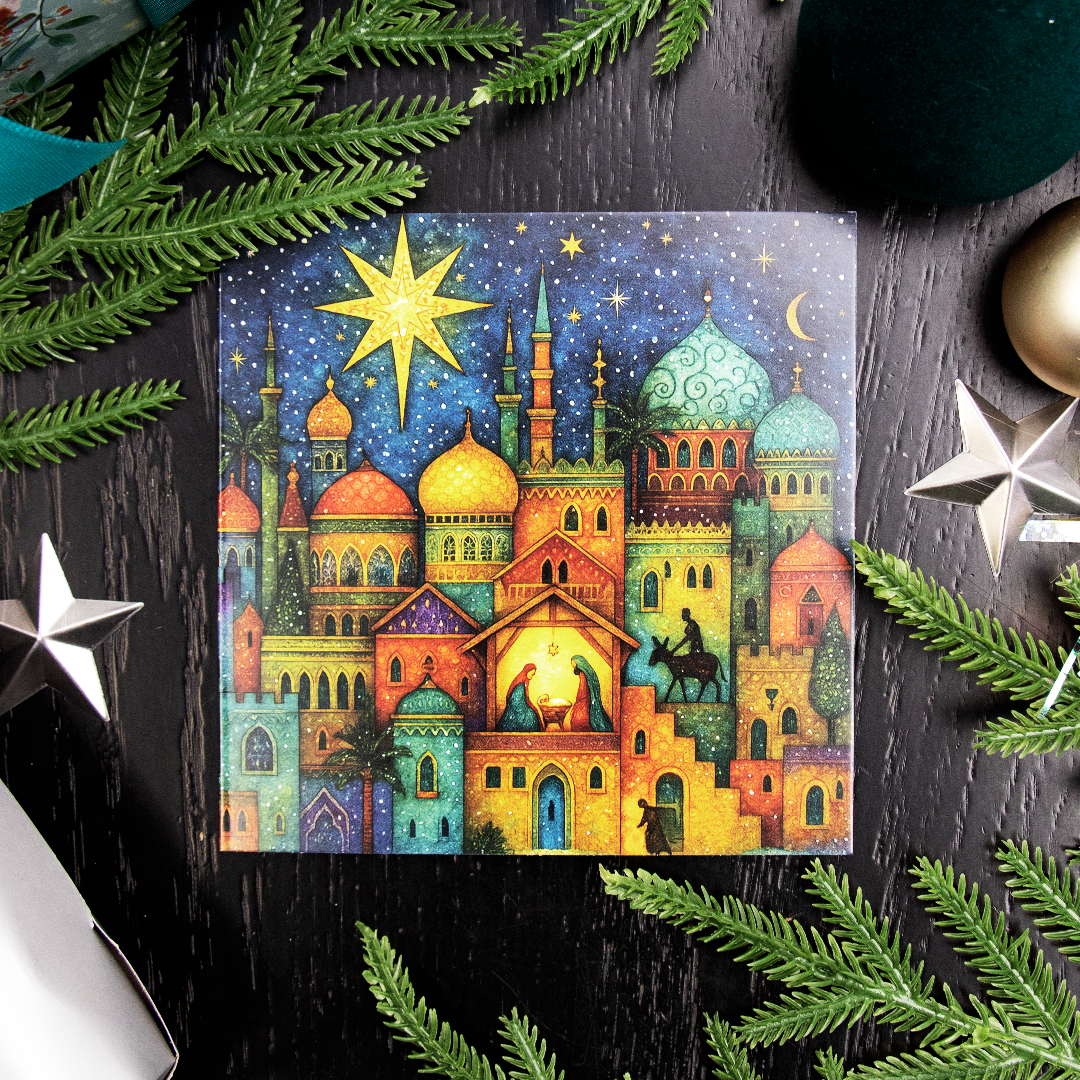 A square Christmas greetings card featuring a colorful Christmas nativity scene in Bethlehem with domed buildings, a glowing star, and a stable under a dark blue sky, styled on a dark wood surface with pine branches and silver star ornaments.