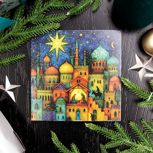 A square Christmas greetings card featuring a colorful Christmas nativity scene in Bethlehem with domed buildings, a glowing star, and a stable under a dark blue sky, styled on a dark wood surface with pine branches and silver star ornaments.