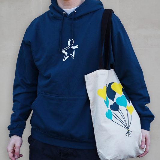 A model is shown from just below the head to the torso against a grey background. The model wears a blue hoodie with a front pocket, drawstrings, and a white Star of Hope printed on the chest. A tote bag hangs over their left shoulder, featuring dark teal, light teal, and yellow heart‑shaped balloon designs on the front.