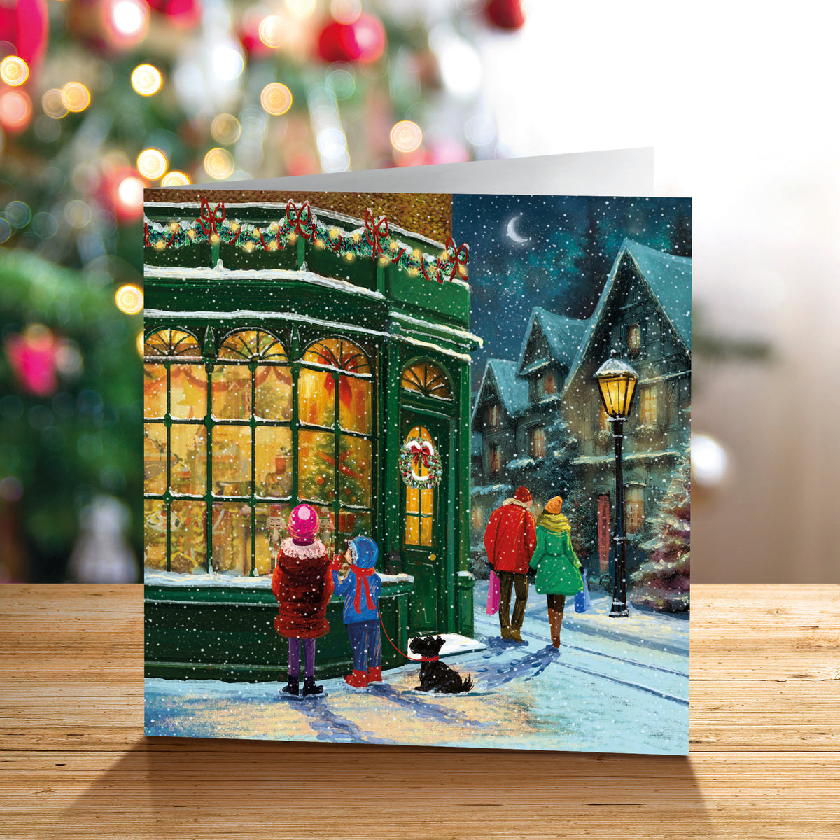 A square Christmas greeting card standing on a wooden surface, featuring a snowy village street scene with a green shop, glowing windows, people walking, and a dog. The background is a softly blurred festive holiday setting with a Christmas tree.