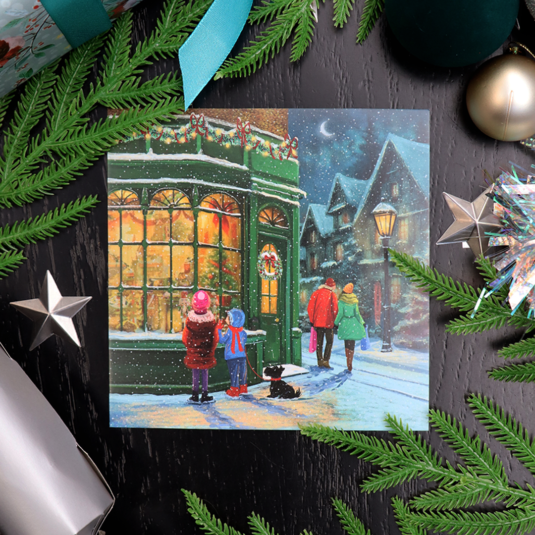 A square Christmas card featuring a snowy village street scene with children watching a green storefront, surrounded by festive green pine branches, ornaments, and gift wrapping on a dark black wood surface.