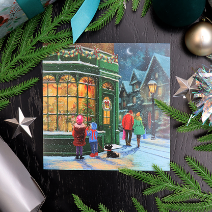 A square Christmas card featuring a snowy village street scene with children watching a green storefront, surrounded by festive green pine branches, ornaments, and gift wrapping on a dark black wood surface.