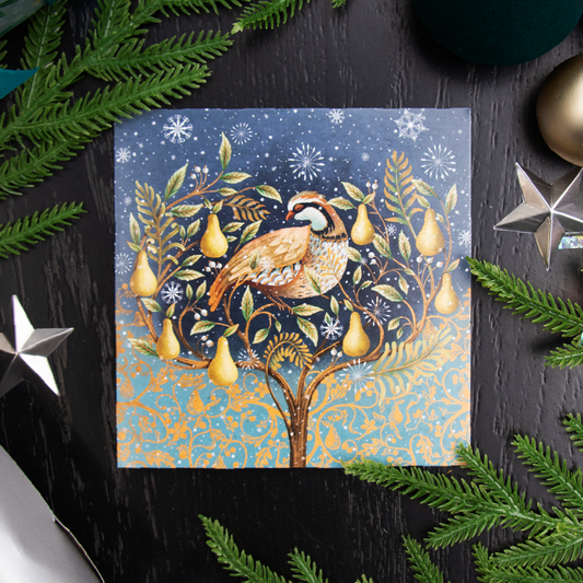 A square Christmas greetings card or featuring a partridge bird perched on a pear tree with snowflakes, styled on a dark wood surface with green pine branches and silver star ornaments around it.