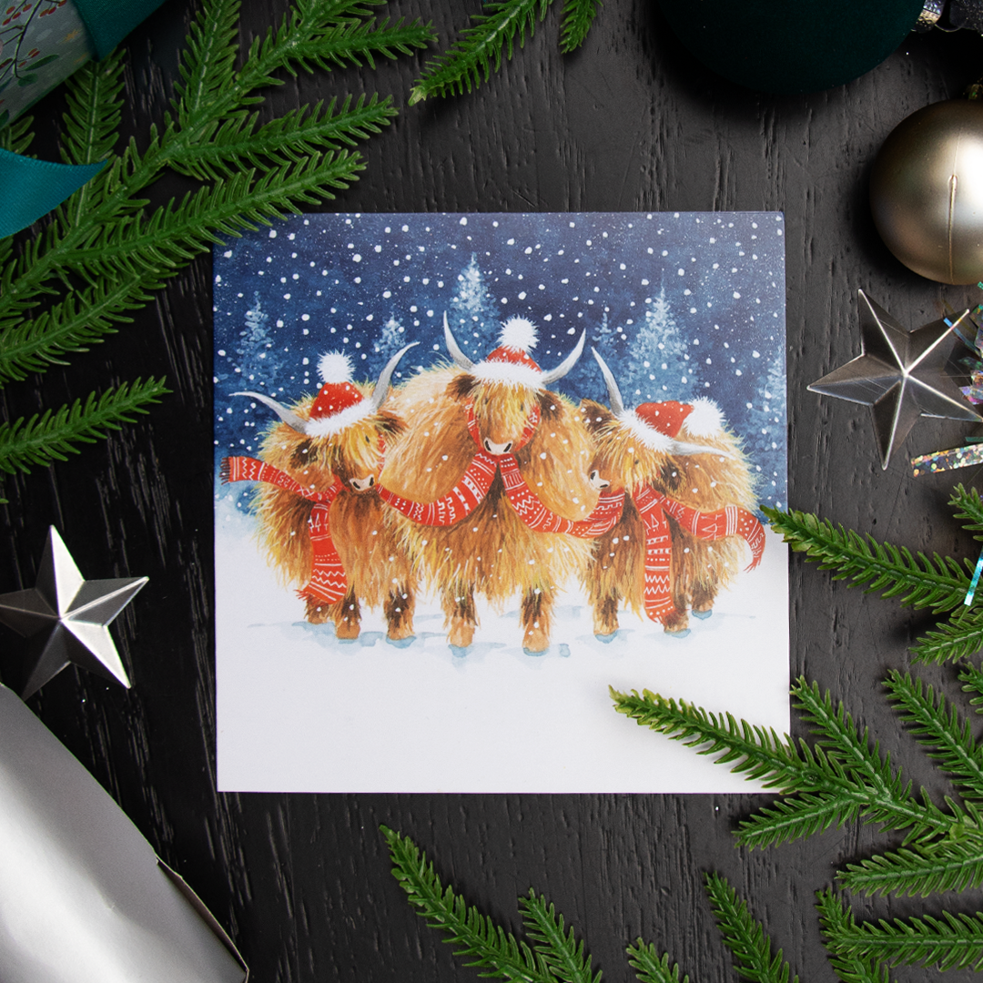 A square Christmas greetings card featuring an illustration of three Highland cows wearing red Santa hats and scarves under falling snow, styled in a flat lay with green pine branches and holiday ornaments on a dark wood surface.