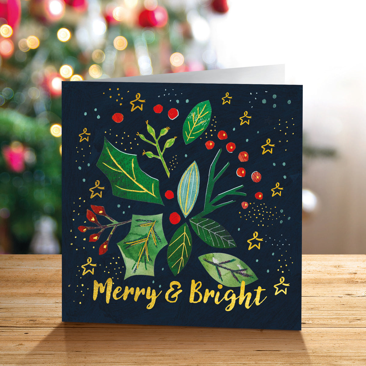 A square Christmas greeting card standing on a wooden surface, featuring a dark navy background with green leaves, red berries, gold Stars of Hope, and the words 'Merry & Bright' in gold. The card is photographed in front of a softly blurred Christmas tree with warm lights.