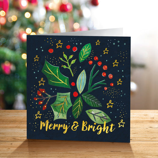 A square Christmas greeting card standing on a wooden surface, featuring a dark navy background with green leaves, red berries, gold Stars of Hope, and the words 'Merry & Bright' in gold. The card is photographed in front of a softly blurred Christmas tree with warm lights.
