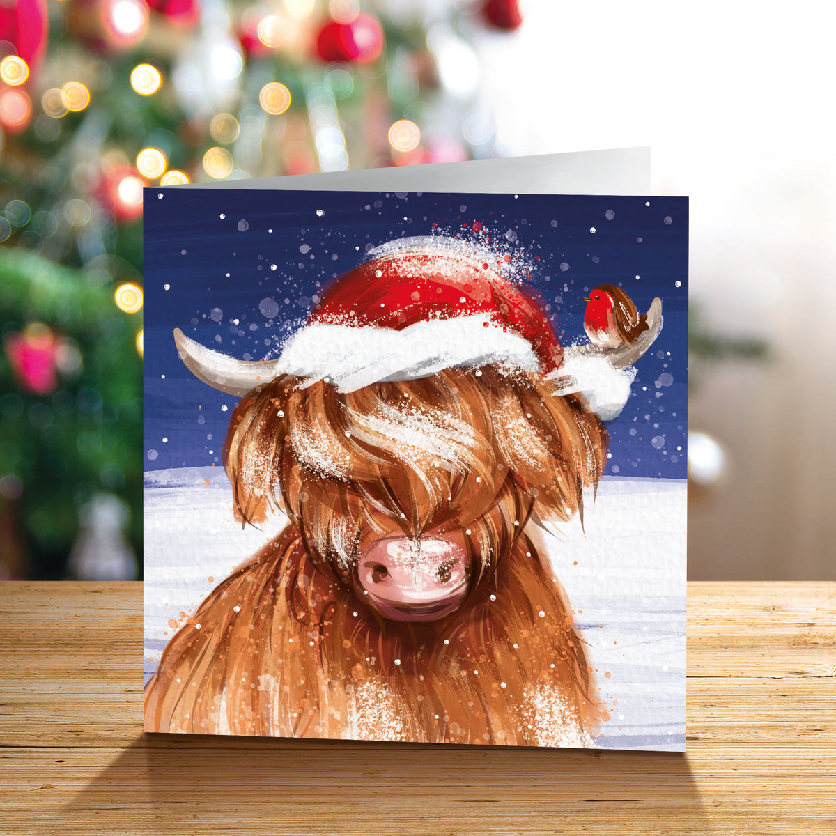 A square Christmas card featuring a Highland cow wearing a red Santa hat with white trim, standing on a wooden surface with a blurred Christmas tree in the background.