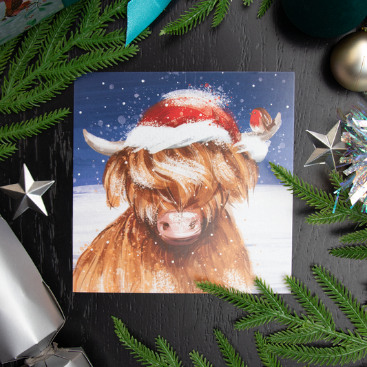 A square Christmas card featuring a Highland cow wearing a Santa hat in the snow, styled in a flat-lay with pine branches, silver ornaments, and a dark black wood background.