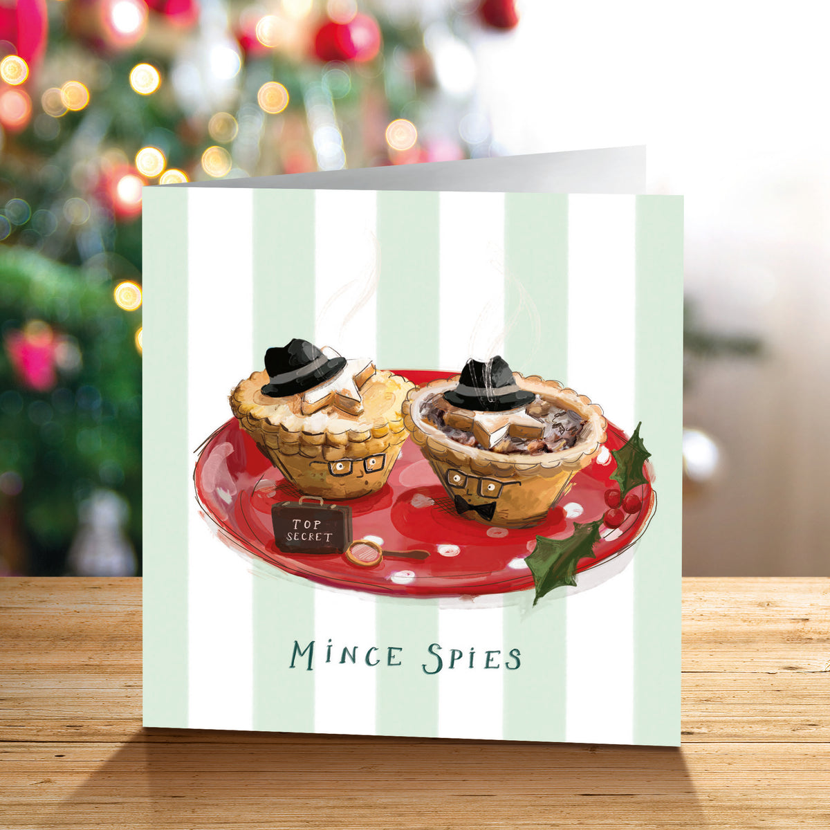 A Christmas greetings card featuring an illustration of two mince pies dressed in black hats and glasses, on a red plate with the text 'Mince Spies' below, displayed on a wooden surface with a blurred Christmas tree in the background.