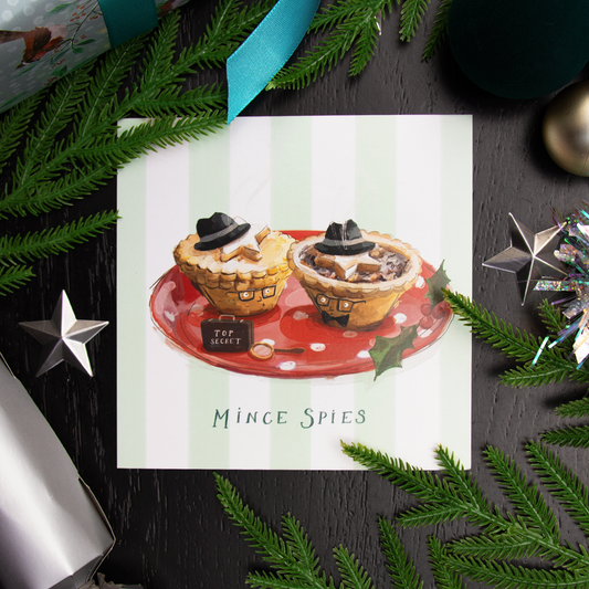 A square Christmas greetings card featuring an illustration of two mince pies wearing hats and glasses, on a red plate, surrounded by green pine branches and holiday decorations on a dark black wooden surface. The card reads 'Mince Spies' at the bottom.