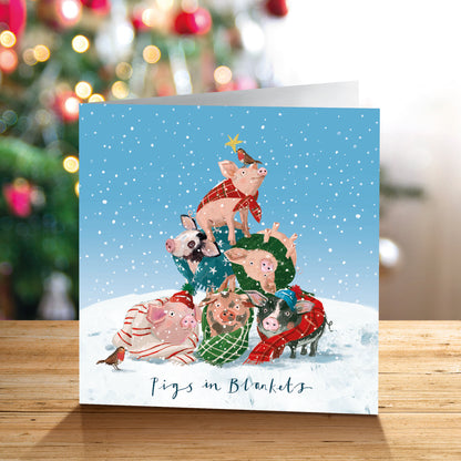 A square Christmas greetings card standing on a wooden surface, featuring an illustration of pigs bundled in winter clothing and blankets under a snowy blue sky. The card reads 'Pigs in Blankets' at the bottom.