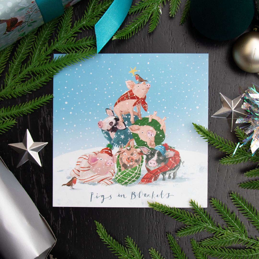 A square Christmas greetings card featuring an illustration of pigs dressed in holiday clothing, stacked together in a winter scene, with the text 'Pigs in Blanket'. The card is styled in a flat-lay on a dark black wood background with pine branches, silver ornaments, and a teal ribbon.