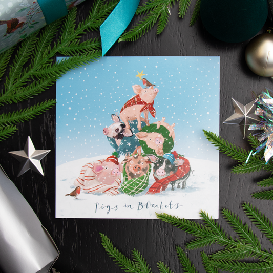 A square Christmas greetings card featuring an illustration of pigs dressed in holiday clothing, stacked together in a winter scene, with the text 'Pigs in Blanket'. The card is styled in a flat-lay on a dark black wood background with pine branches, silver ornaments, and a teal ribbon.