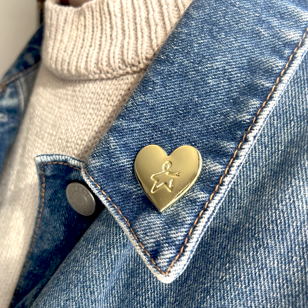 Gold heart shape pin badge attached to the collar of a blue denim jacket worn by a model, featuring an engraved Star of Hope at the centre of the pin badge.