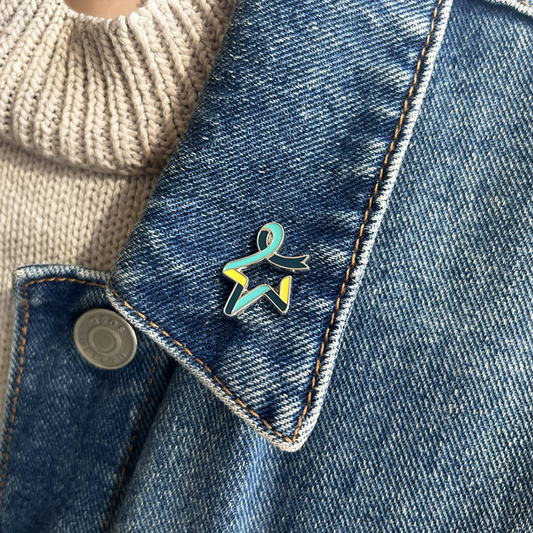 Our Star of Hope pin badge with a dark teal, light teal, and yellow multicoloured Star of Hope design, attached to the collar of a blue denim jacket worn by a model.