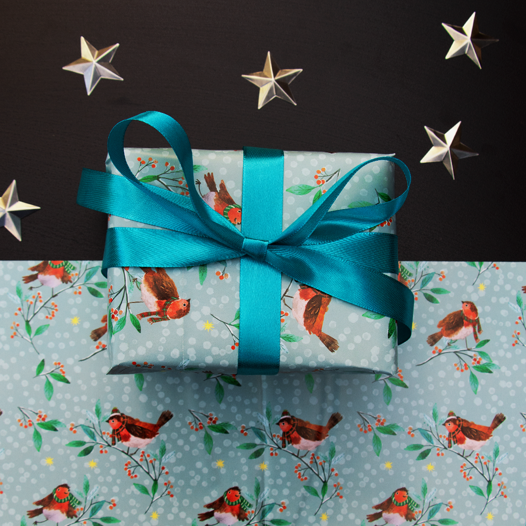 A wrapped gift box tied with a bright teal ribbon, placed on top of matching holiday wrapping paper printed with red birds, green leaves, and small red berries. The scene is styled with metallic star decorations on a dark black background.