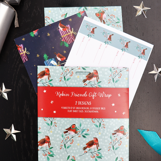 A gift wrap set displayed on a dark black tabletop with decorative star cutouts. The set includes multiple wrapping paper designs featuring robin birds, red berries, and a dark navy holiday pattern, along with white gift tags and a red paper band reading 'Robin Friends Gift Wrap 2 DESIGNS'.