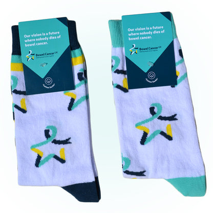 Two pairs of our Star of Hope socks are shown with a dark‑ and light‑teal label attached, featuring the text: “Our vision is a future where nobody dies of bowel cancer.” The pair on the left is white with the Star of Hope logo and dark teal, yellow, and light‑teal stripes around the ankles and feet. The pair on the right is white with the Star of Hope logo running vertically down the sides, and light‑teal toes and ankle cuffs.