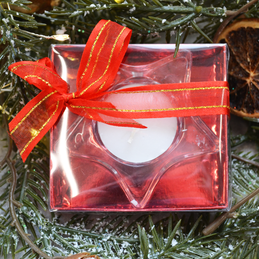 Star-shaped glass candle holder with a white tea light, presented in a clear gift box with a red interior and red-and-gold ribbon, surrounded by pine branches and dried orange slices.