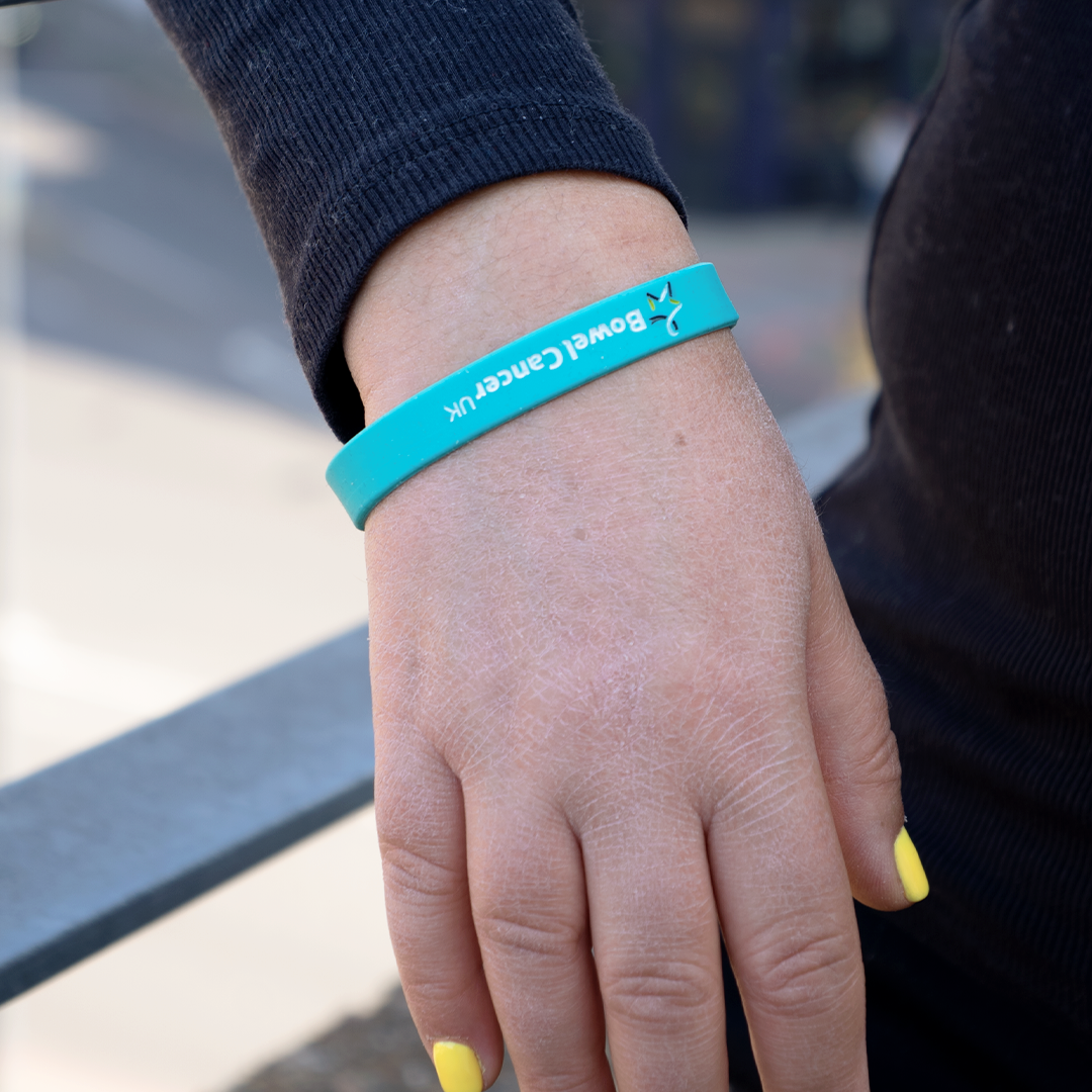 Close‑up of a model’s right hand wearing a teal wristband featuring a small multicoloured Star of Hope and “Bowel Cancer UK” printed in white text.