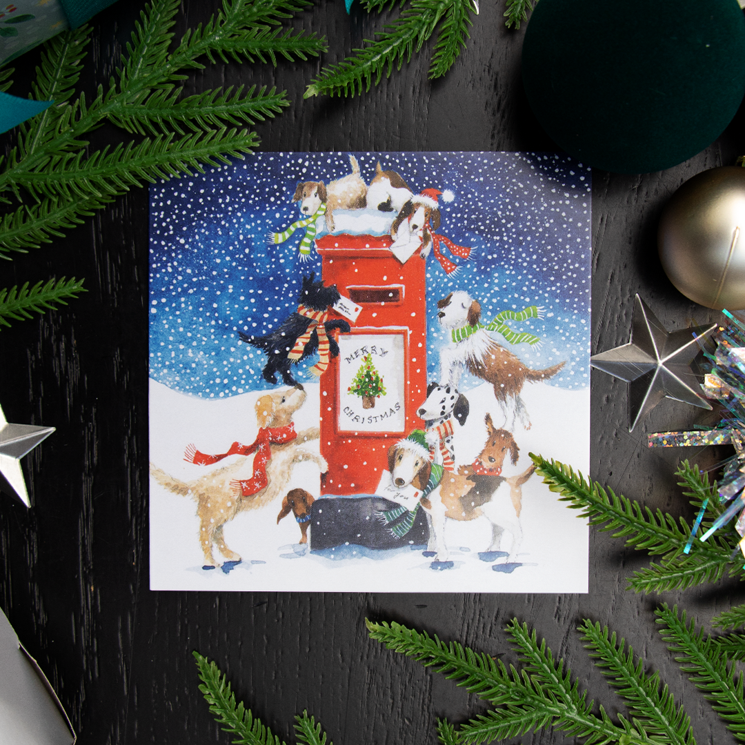 A square Christmas greetings card featuring a festive illustration of dogs around a red post box in the snow, with the text 'MERRY CHRISTMAS' on front of the postbox. The card is styled in a flat-lay on a dark wooden surface surrounded by green pine branches and holiday decorations.