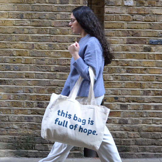 Female model walking right to left in front of a brick wall, carrying the Weekender Tote Bag over their left arm. The front of the bag is visible, printed with the message “this bag is full of hope” in dark teal text.