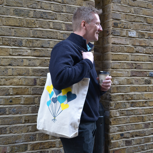 Male model standing in front of a brick wall, facing right. The model carries a Balloon Heart tote bag over their right shoulder, featuring yellow, dark teal, and light teal balloon‑shaped hearts printed on the front of the bag. The model also holds a white disposable coffee cup with a black lid in their left hand.