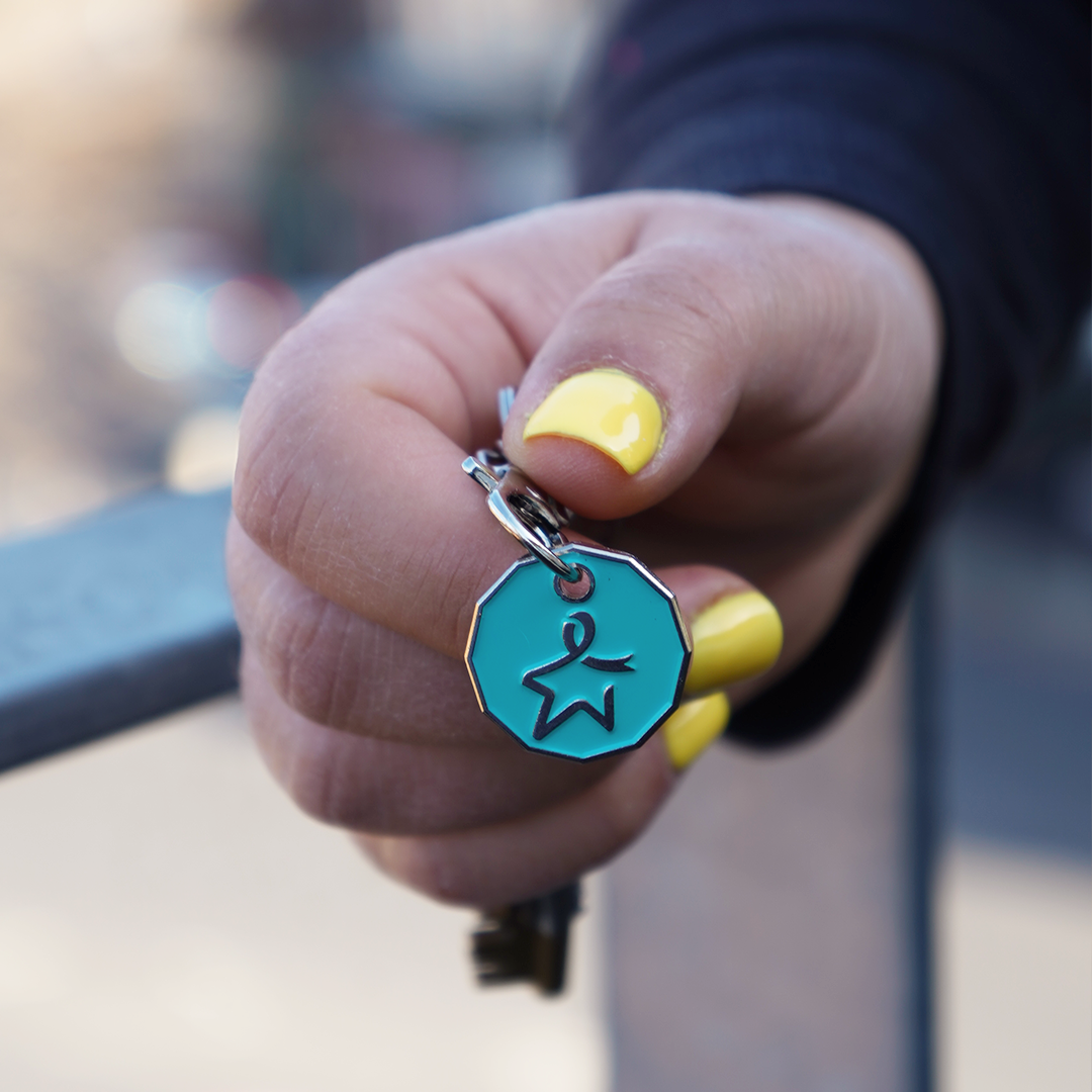 Close‑up of a model’s right hand holding a set of keys with a teal trolley coin featuring an engraved Star of Hope.