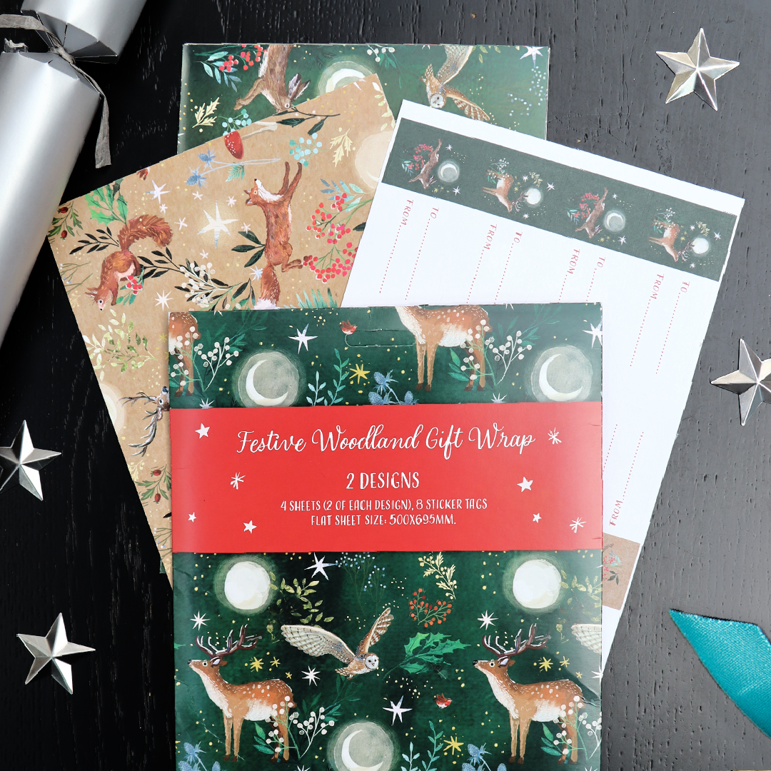Festive woodland gift wrap set displayed on a dark black wooden surface with silver star decorations and a silver ribbon. The set includes a green wrap with deer, owls, moons and stars, as well as a tan/brown wrap with woodland animals, and a white sheet with printed labels.
