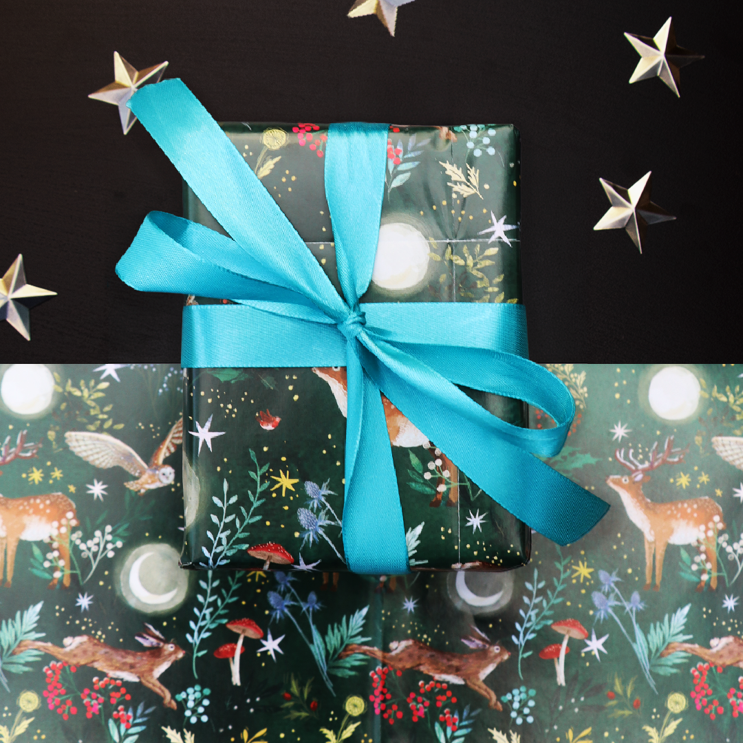 A wrapped gift box tied with a teal ribbon, placed on top of matching festive wrapping paper with a pattern featuring deers, hares, owls, moons and foliage. The background is black with scattered metallic star decorations.