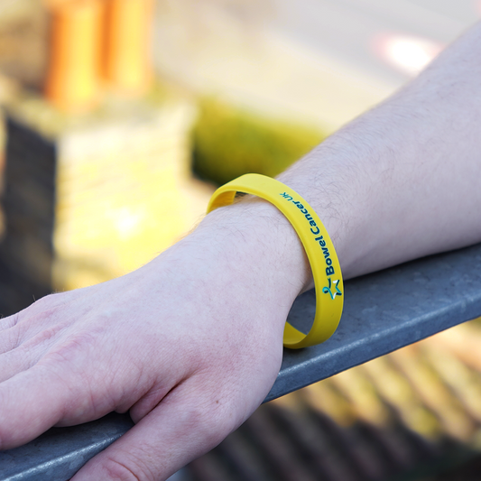 Close‑up of a model’s right hand resting on a grey metal railing, wearing a yellow wristband featuring a small multicoloured Star of Hope and “Bowel Cancer UK” in dark teal text.